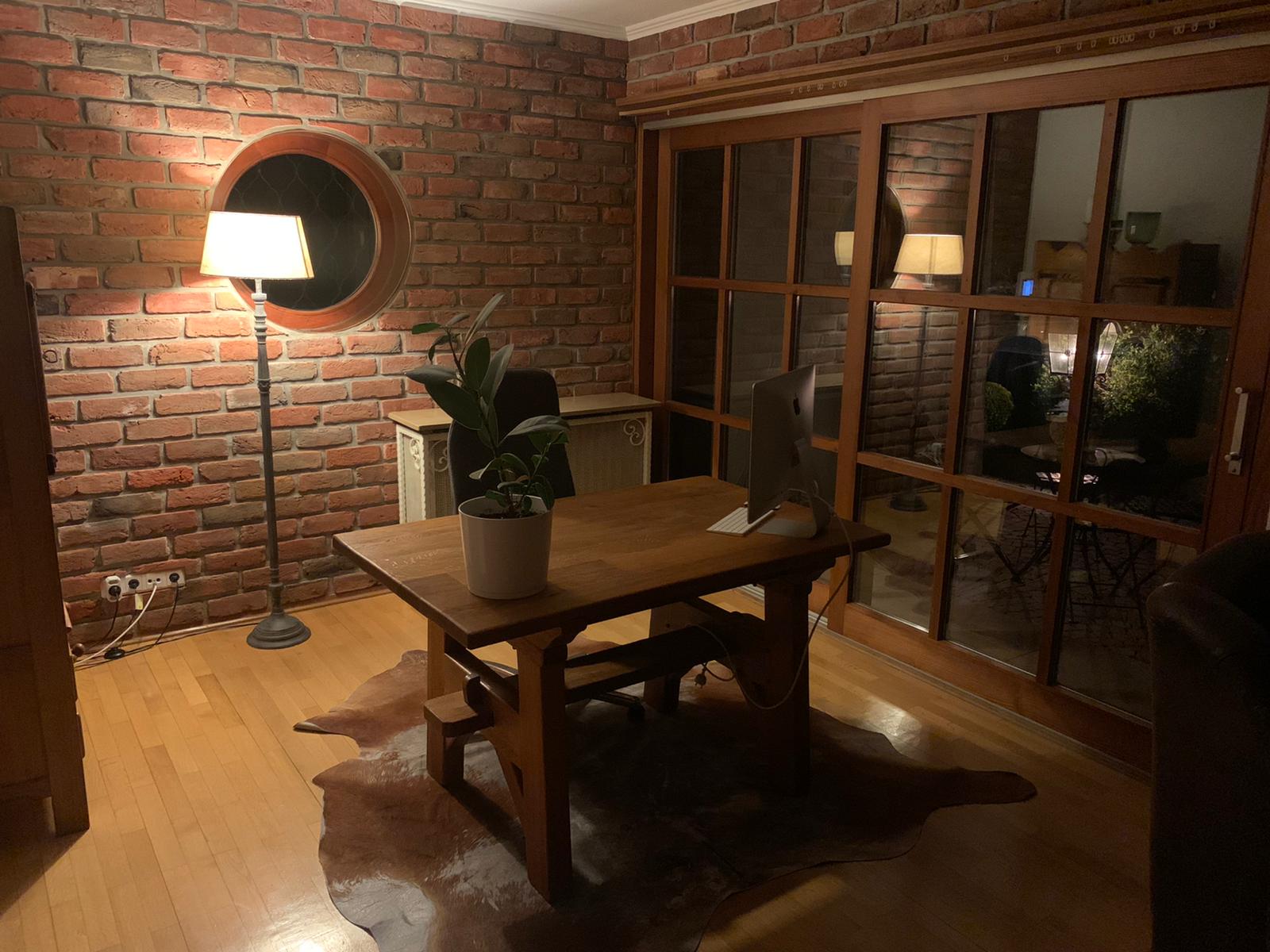 Stimmungsvolles Homeoffice mit rustikaler Einrichtung und einer warmtonigen Wandverkleidung aus recycelten Backsteinverblendern. Die runde Fensteröffnung und das warme Licht verleihen dem Raum eine einladende, kreative Atmosphäre.