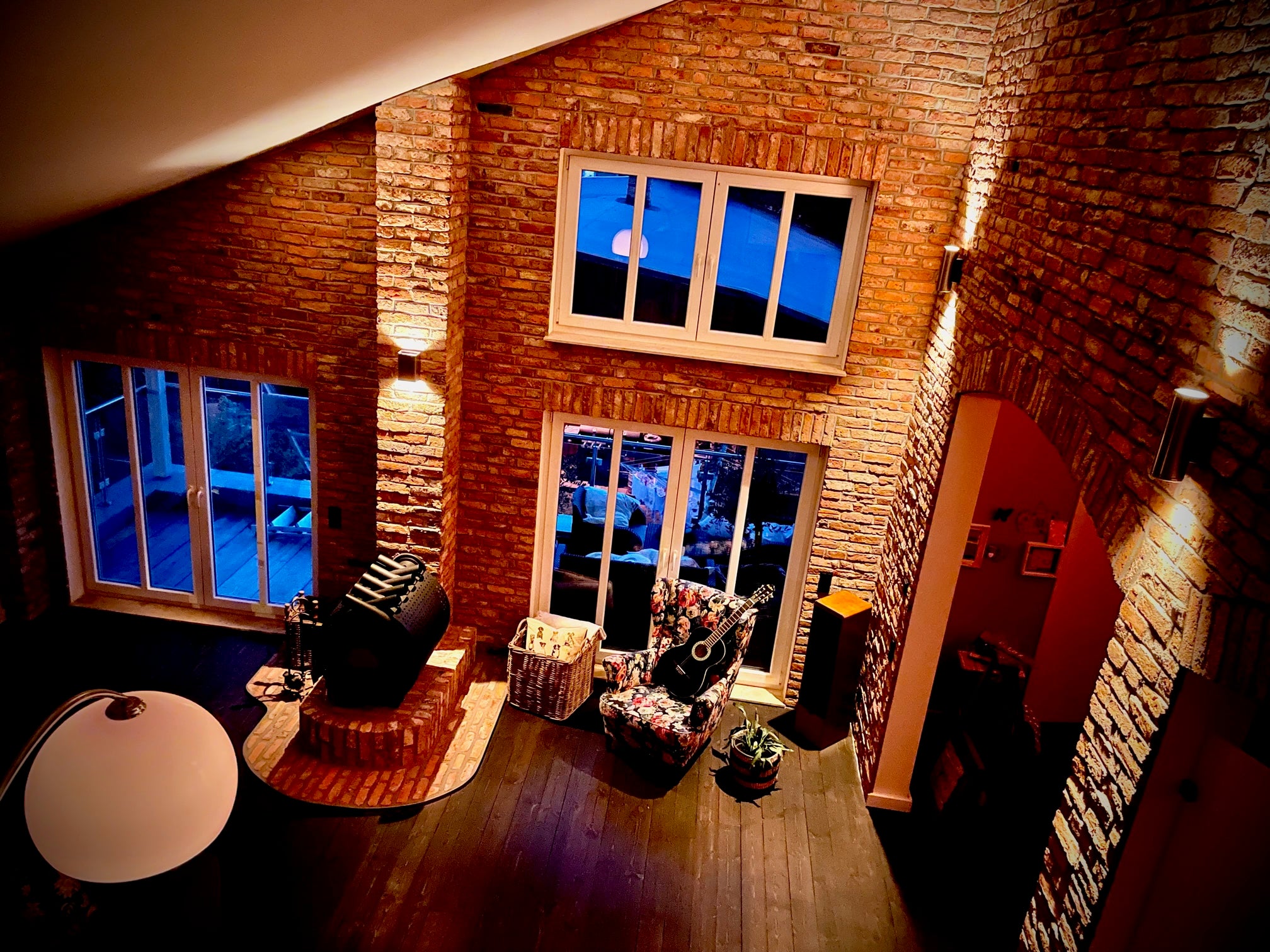 Großzügiges Wohnzimmer mit offener Galerie, verkleidet mit wiederverwendetem Sichtmauerwerk aus recycelten Backsteinriemchen. Die Kombination aus Ziegelsäulen, Kaminofen und Lichtakzenten schafft ein stilvolles Ambiente mit Loft-Charakter.