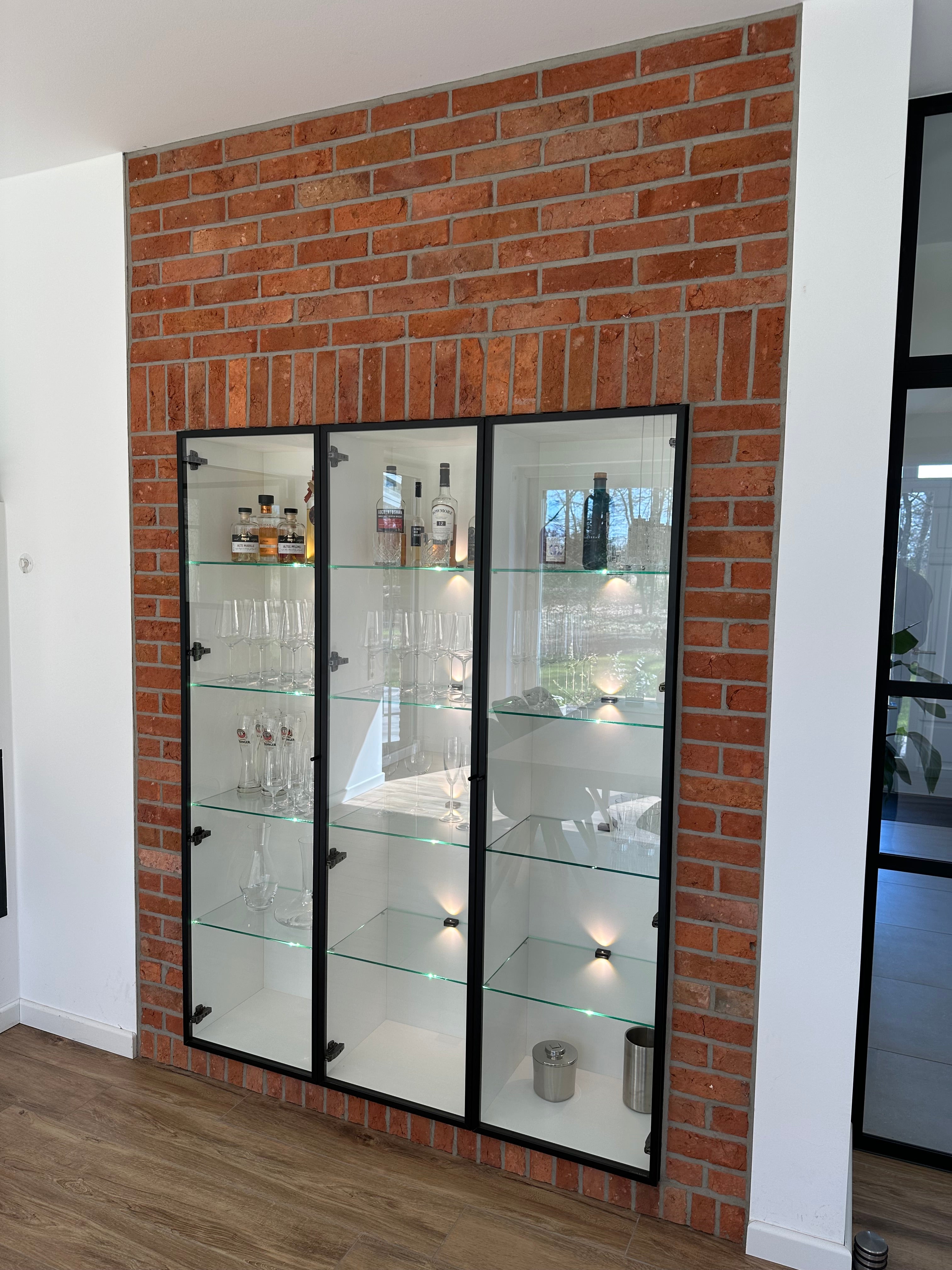 Stilvolles Wohnzimmer mit moderner Glasvitrine vor einer Ziegelwand aus recyceltem Backstein, akzentuiert durch gezielte Beleuchtung. Die Ziegelstruktur sorgt für eine gemütliche, rustikale Atmosphäre, die perfekt mit der klaren Form der Vitrine harmoniert.