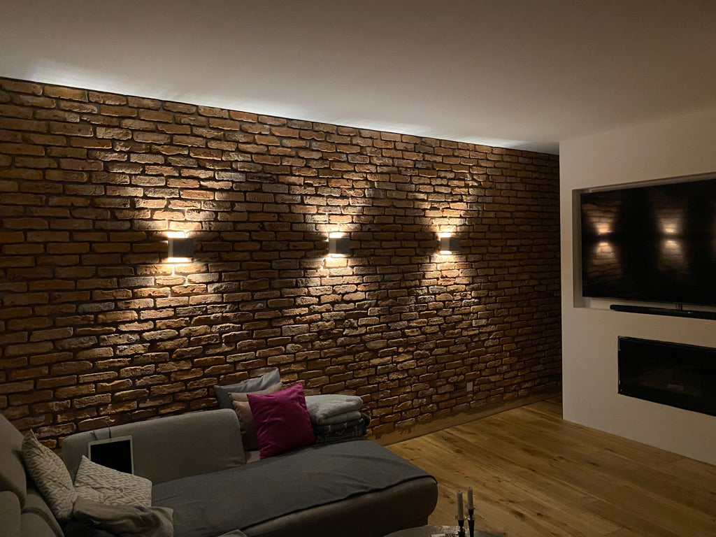 Elegantes Wohnzimmer mit strukturierter Ziegelwand aus recycelten Altziegeln, akzentuiert durch indirekte Wandleuchten, TV-Fläche und integriertem Kamin – harmonische Kombination aus Technik und Handwerk