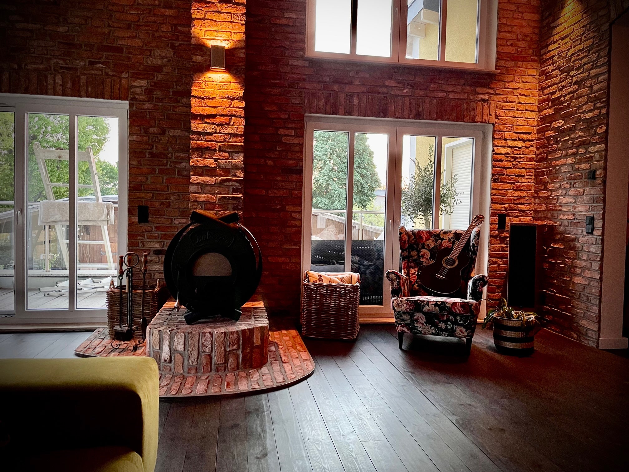 Lichtdurchflutetes Wohnzimmer mit einem freistehenden Kaminofen und detailreicher Raumverkleidung aus wiederverwendetem Mauerziegel. Die sichtbare Ziegelstruktur verleiht dem Raum eine warme, handwerkliche Atmosphäre mit ländlichem Charme.