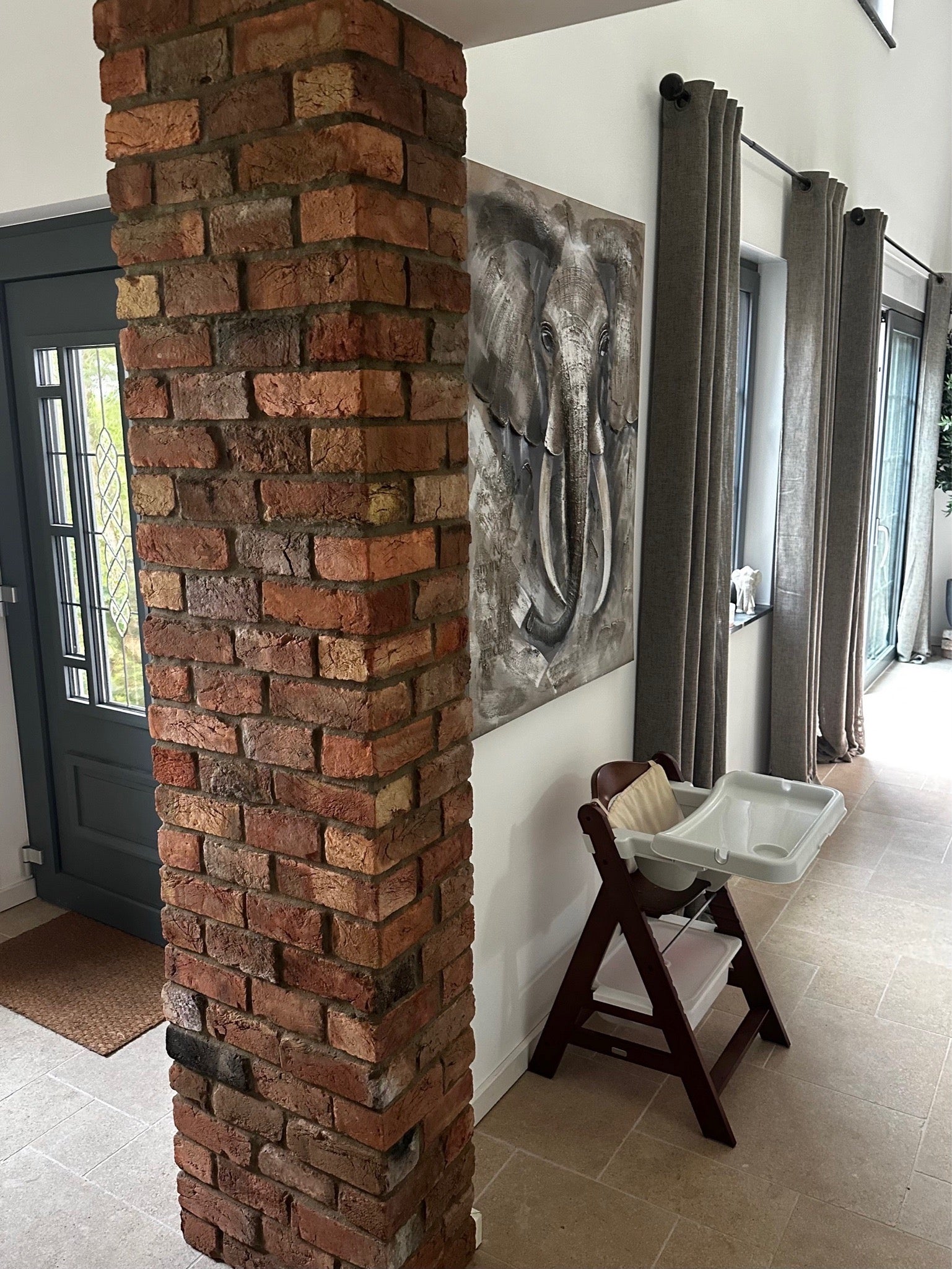 Lichtdurchfluteter Wohnbereich mit markanter Ziegelsäule aus recycelten Altziegeln, kombiniert mit modernen Elementen, Kinderstuhl und Tierkunst – harmonische Mischung aus Funktionalität und Charakter